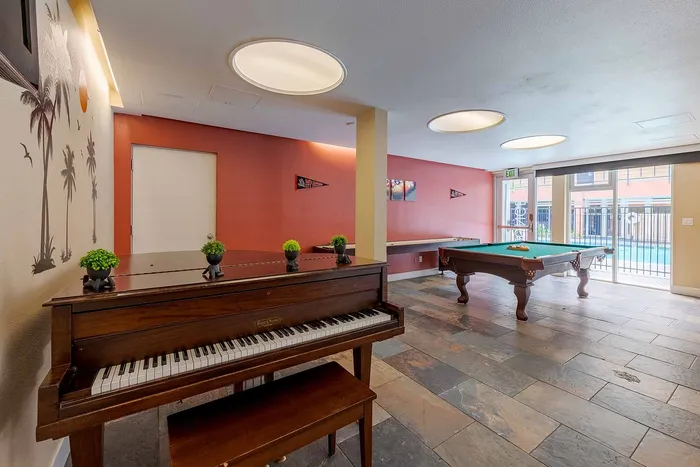 Elegant recreation room with a classic wooden piano, vibrant wall art, and a stylish pool table overlooking a refreshing pool area.