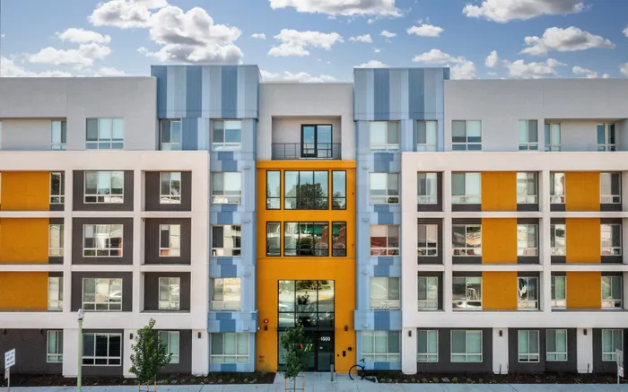 Vibrant modern apartment building with colorful facade and large windows, set against a bright blue sky with fluffy clouds.