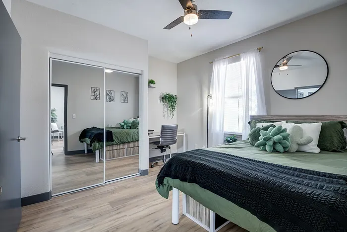 Inviting bedroom with chic decor, featuring a plush bed with green accents, mirrored closet doors, and ample natural light from a large window.