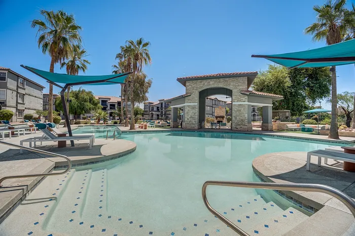 Luxurious swimming pool area with shaded loungers surrounded by lush palm trees and modern apartment buildings under a clear blue sky.