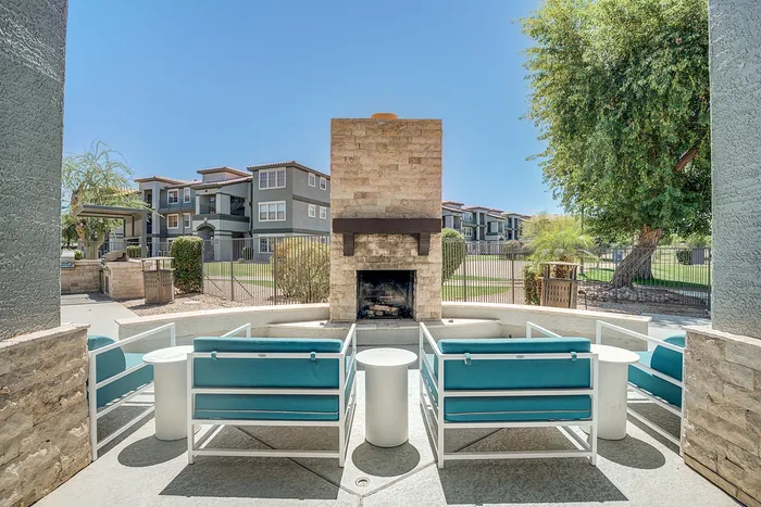 Chic outdoor lounge area with modern seating and a stone fireplace, surrounded by lush greenery and elegant apartment buildings in the background.