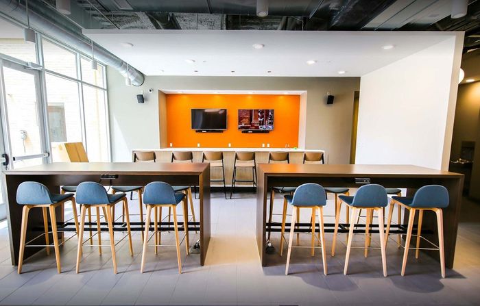 Contemporary communal lounge with vibrant orange accent wall, stylish bar seating, and ample natural light from large windows.