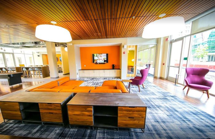 Modern and vibrant living space featuring a stylish orange sofa, elegant purple chairs, and a sleek entertainment center, illuminated by large windows and contemporary lighting.