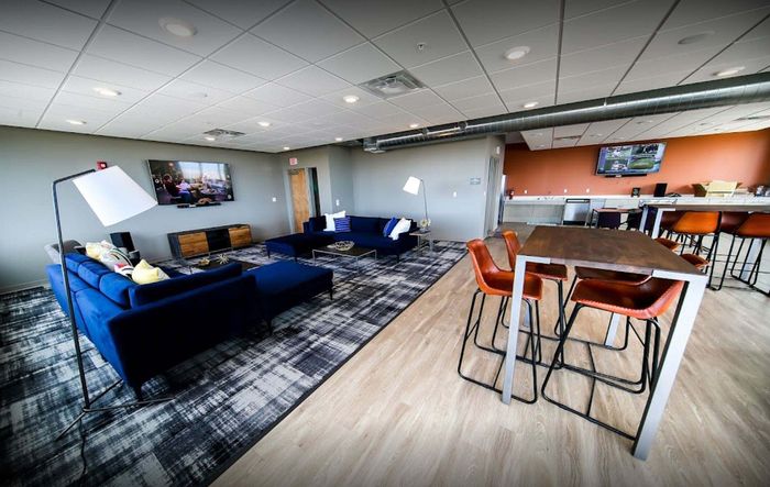 Modern apartment lounge with chic blue sofas, vibrant decor, and a sleek open-concept layout featuring bar seating and a large wall-mounted TV.