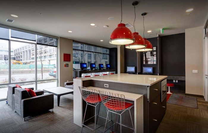 Modern apartment community space featuring a spacious layout with stylish red pendant lights, sleek countertops, and comfortable seating with a vibrant view of the city through large windows.