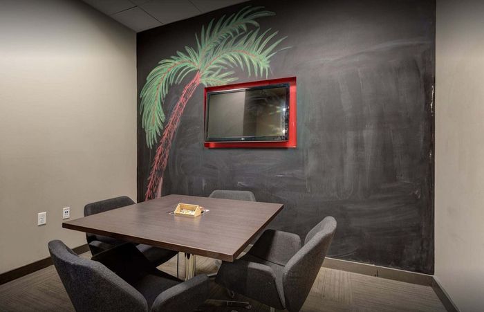 Modern meeting room with a sleek table, comfortable chairs, a wall-mounted TV, and a chalkboard wall featuring a vibrant palm tree drawing.