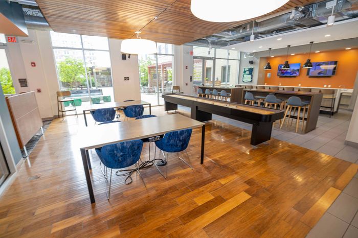 Modern communal space with polished wooden floors, vibrant seating, a shuffleboard table, and a stylish bar area with multiple screens.