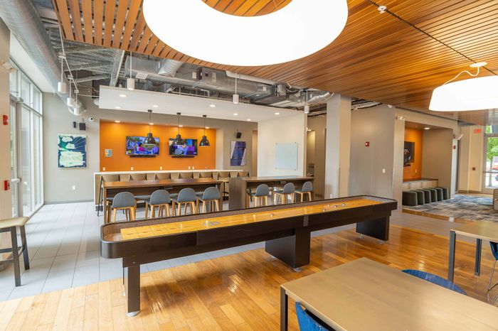 Modern communal lounge with a sleek shuffleboard table, multiple seating areas, vibrant decor, and large windows for natural light.