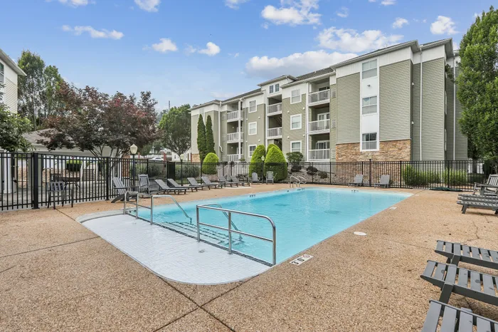 Inviting apartment complex featuring a sparkling pool surrounded by lounge chairs, lush landscaping, and modern multi-story buildings under a bright blue sky.