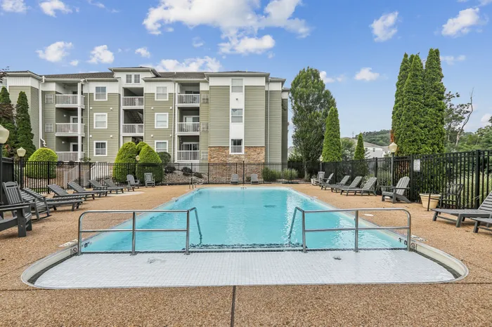 Elegant apartment complex with a pristine swimming pool, surrounded by lush greenery and comfortable loungers, set against a bright, blue sky.