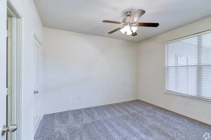 Bright and airy room with plush carpeting, featuring a modern ceiling fan and large window for natural light.
