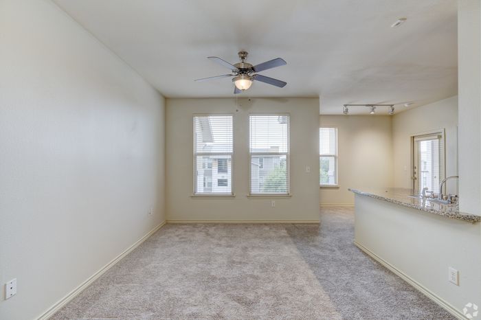 Bright and airy apartment living room with plush carpeting, large windows for natural light, and a modern ceiling fan next to a sleek granite kitchen counter.