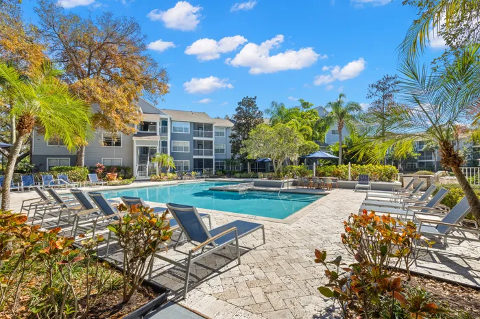Outdoor pool with tiled flooring, seating, and surrounding trees.