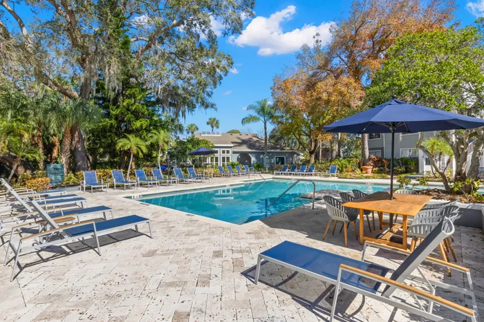 Outdoor pool with tiled flooring, seating, and surrounding trees.