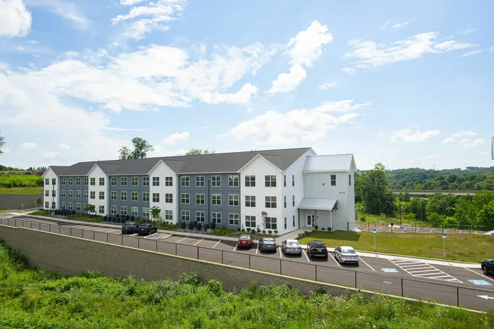 Modern apartment building with stylish architecture, ample parking, surrounded by lush greenery and a clear blue sky.