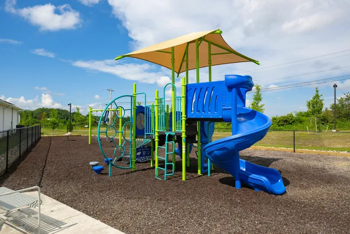 Vibrant outdoor playground with a blue slide and climbing structures under a sunshade, surrounded by lush greenery and open skies.