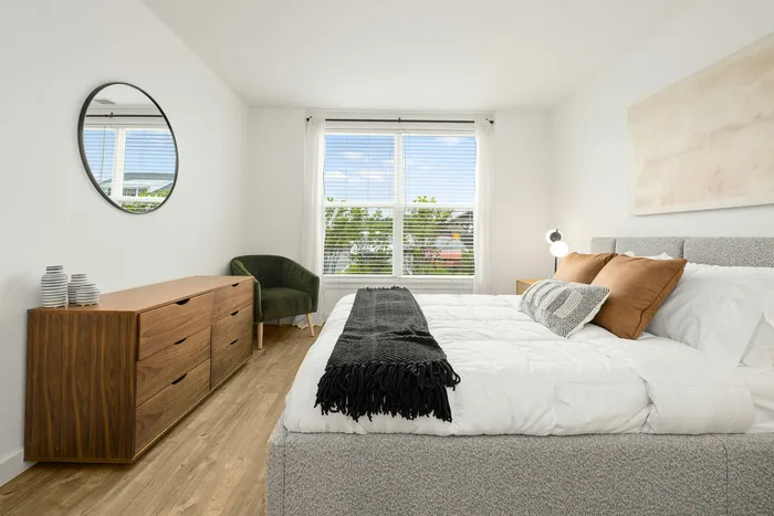 Bright and inviting bedroom with sleek modern decor, featuring a large window, cozy bed, stylish wooden dresser, and elegant green accent chair.