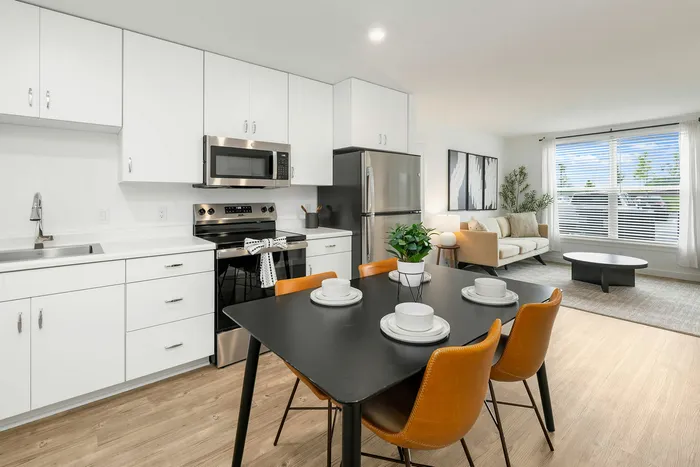Modern open-plan apartment featuring sleek white kitchen with stainless steel appliances, a stylish dining area with leather chairs, and a cozy living room with large windows.