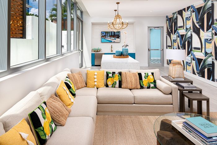 Vibrant living room featuring a modern L-shaped sofa with colorful pillows, large windows allowing natural light, and a stylish chandelier above a sleek kitchen island.