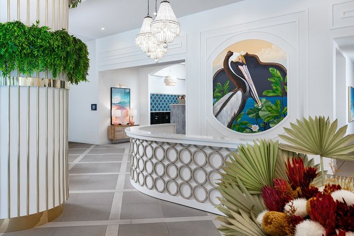 Elegant lobby with modern decor, featuring a stunning chandelier, vibrant artwork of a pelican, and lush greenery, creating a welcoming atmosphere.