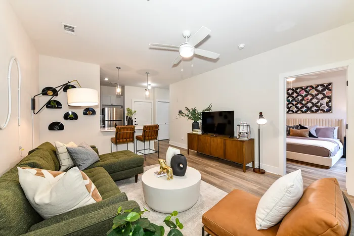 Inviting open-plan living space with modern decor, featuring a cozy living room with a stylish green sofa and chic wall art, adjacent to a sleek kitchen with bar seating, and a glimpse of a comfortable bedroom.