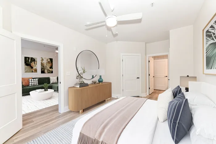 Inviting bedroom with modern decor, featuring elegant furnishings, a stylish mirror, and a glimpse into a cozy living room with chic artwork and a plush green sofa.