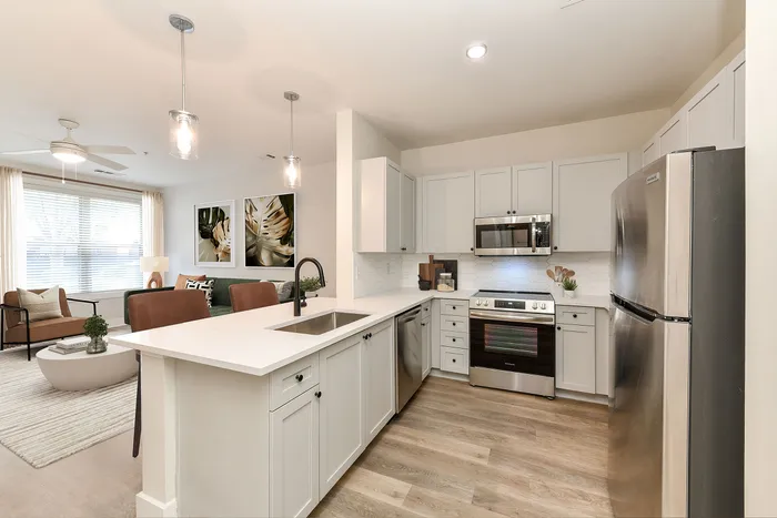 Contemporary open-concept kitchen and living area featuring sleek white cabinetry, stainless steel appliances, and elegant pendant lighting, with a bright, inviting living room perfect for relaxation.