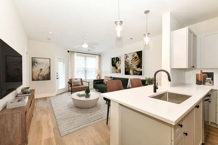 Modern and inviting living space featuring a sleek kitchen with white countertops, cozy seating area, and stylish decor. Natural light pours in through large windows, highlighting the elegant design and warm wooden flooring.