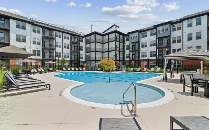 Modern apartment complex featuring a pristine pool with stylish loungers, surrounded by elegant multi-story buildings and lush landscaping.
