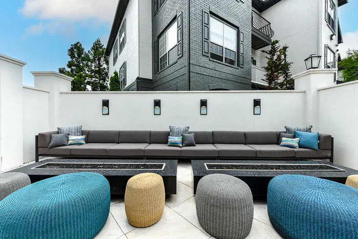 Chic outdoor patio featuring a modern sectional sofa and colorful woven poufs, set against a stylish white and black brick apartment backdrop.