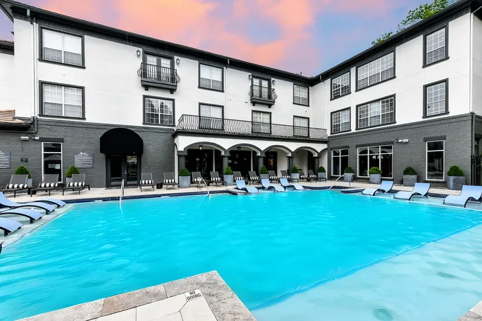 Luxurious apartment complex with a pristine pool surrounded by stylish lounge chairs, set against a backdrop of modern architecture and a stunning sunset sky.