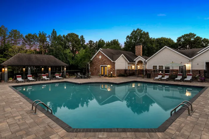 Inviting residential pool area with elegant lounge chairs, surrounded by lush greenery and a charming clubhouse under a twilight sky.