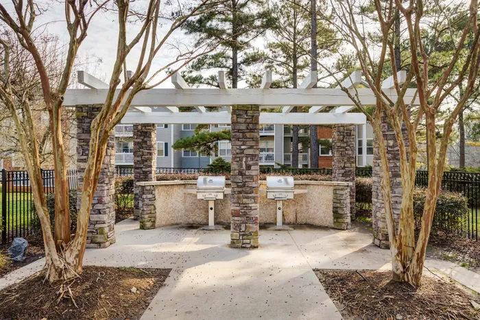 Charming outdoor patio with stone columns and built-in grills, surrounded by lush trees for a perfect barbecue setting.