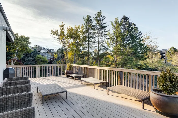 Expansive wooden deck with modern wicker furniture, surrounded by lush greenery and a serene view of trees and neighboring homes.