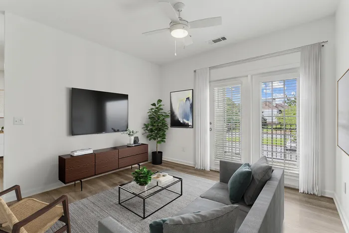 Modern living room with sleek furnishing, featuring a stylish TV setup, cozy seating, and vibrant natural light from large windows overlooking a lush outdoor view.
