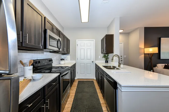 Modern kitchen design with sleek dark cabinets, stainless steel appliances, and elegant white countertops, leading to a cozy, well-lit living area.