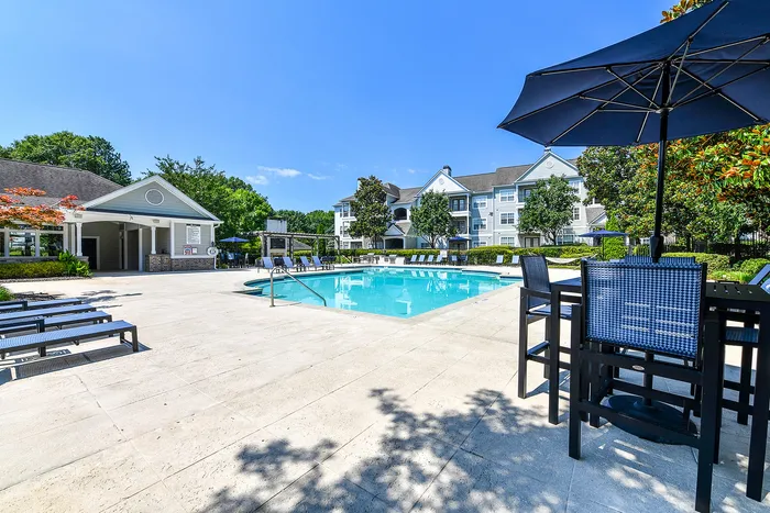 Luxurious apartment complex showcasing a pristine swimming pool, surrounded by elegant lounge chairs and lush greenery, under a clear blue sky.