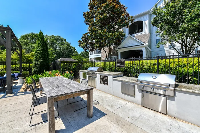 Charming outdoor grilling area with sleek stainless steel barbecues and rustic wooden dining table, surrounded by lush greenery and modern apartments.