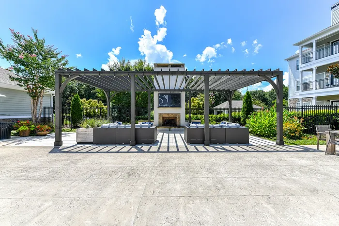 Elegant outdoor patio with pergola, comfortable seating, and fireplace, surrounded by lush gardens under a bright blue sky.