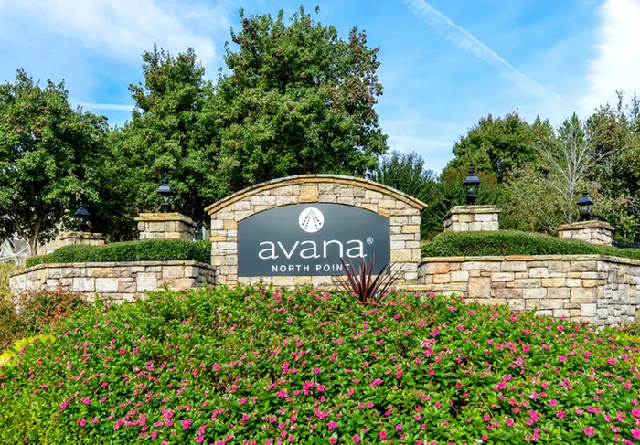 Charming entrance to Avana North Point, featuring a stone wall adorned with vibrant pink flowers and lush greenery, surrounded by serene trees under a bright blue sky.