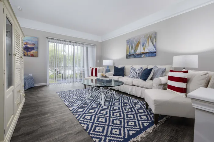 Bright and inviting living room with modern decor, featuring a cozy white sofa, nautical-themed pillows, and a sleek glass coffee table on a stylish geometric rug.