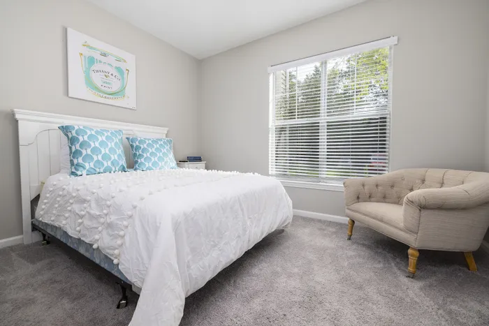 Bright and cozy bedroom featuring a plush bed with blue and white accents, elegant wall art, and a comfortable beige armchair by the window.