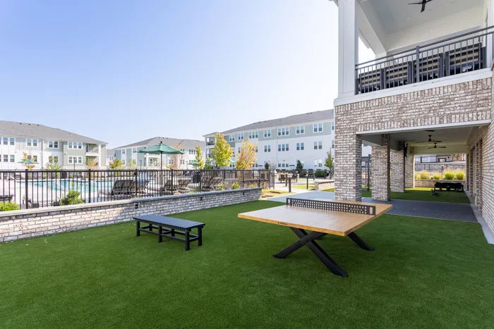 Inviting apartment community featuring a spacious outdoor area with a modern ping pong table, lush green lawn, and a sparkling pool surrounded by comfortable lounge chairs.