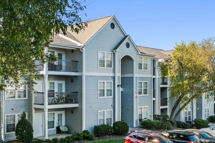 Charming light gray apartment building with inviting balconies, surrounded by lush greenery and ample parking under a clear blue sky.