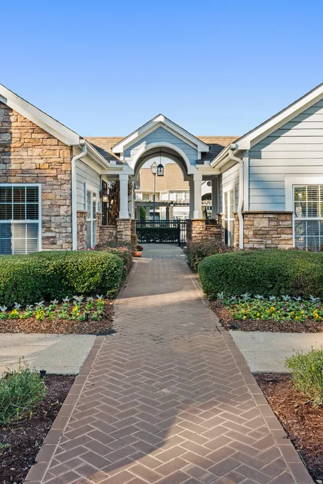 Charming apartment entrance with a welcoming brick pathway, lush landscaping, and elegant stone pillars under a bright blue sky.