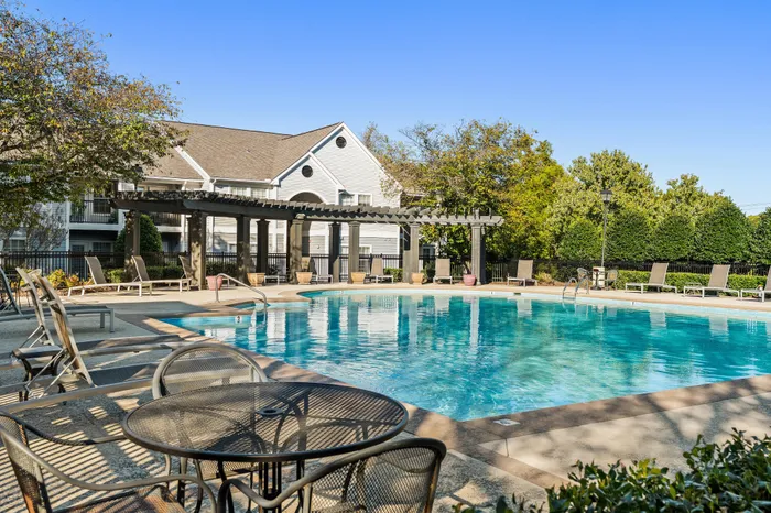 Inviting outdoor pool area with modern lounge chairs and pergola, surrounded by lush greenery and charming apartment homes.
