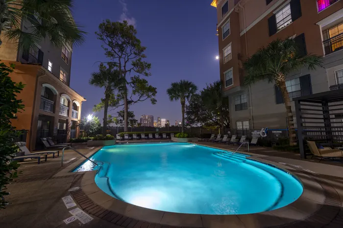 Luxurious apartment poolside view at dusk, featuring serene water, elegant poolside seating, and palm trees with a city skyline backdrop.
