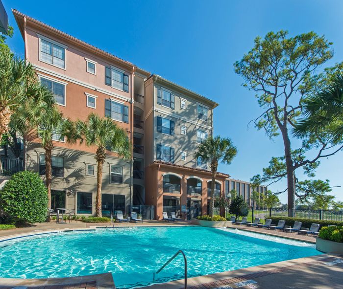 Luxurious apartment complex with a shimmering pool, surrounded by lush palm trees and vibrant landscaping in a sunny setting.