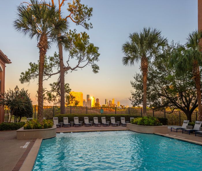Luxurious apartment pool with stunning city skyline views at sunset, surrounded by lush palm trees and comfortable lounging chairs.