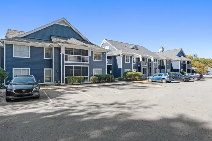 Charming blue apartment complex with modern architecture, ample parking, and lush greenery on a sunny day.